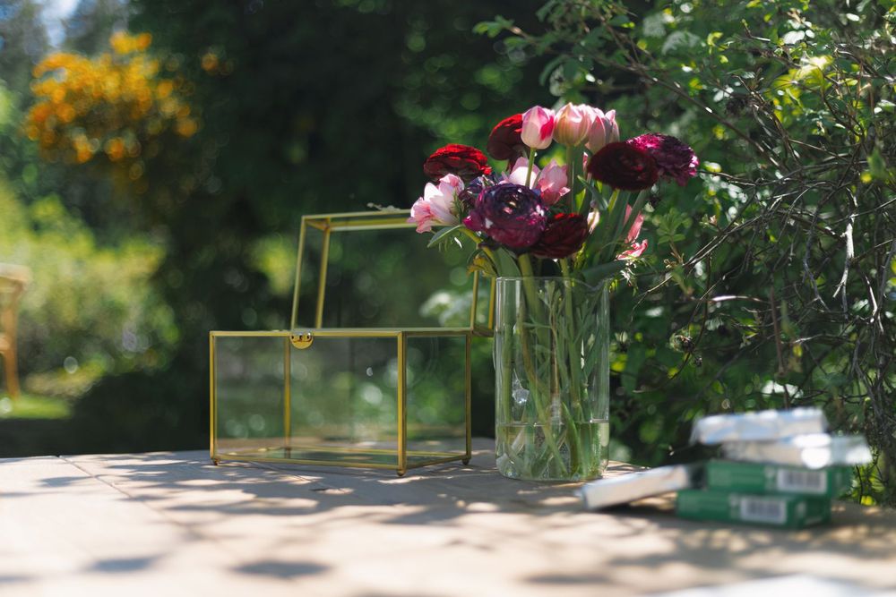 Gold geometric glass boxes and a vase of burgundy and pink ranunculus flowers displayed on an outdoor table.