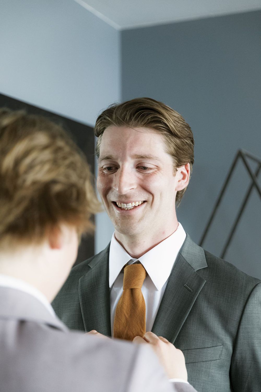 Smiling groom in gray suit and orange tie getting dressed with help from groomsman.