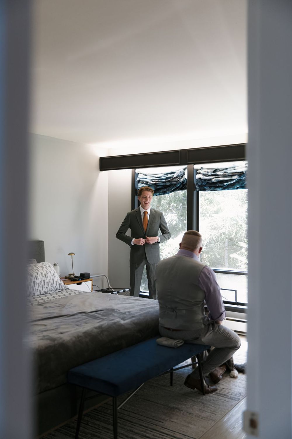 Groom in gray suit prepares in modern bedroom with groomsman seated on bench nearby.