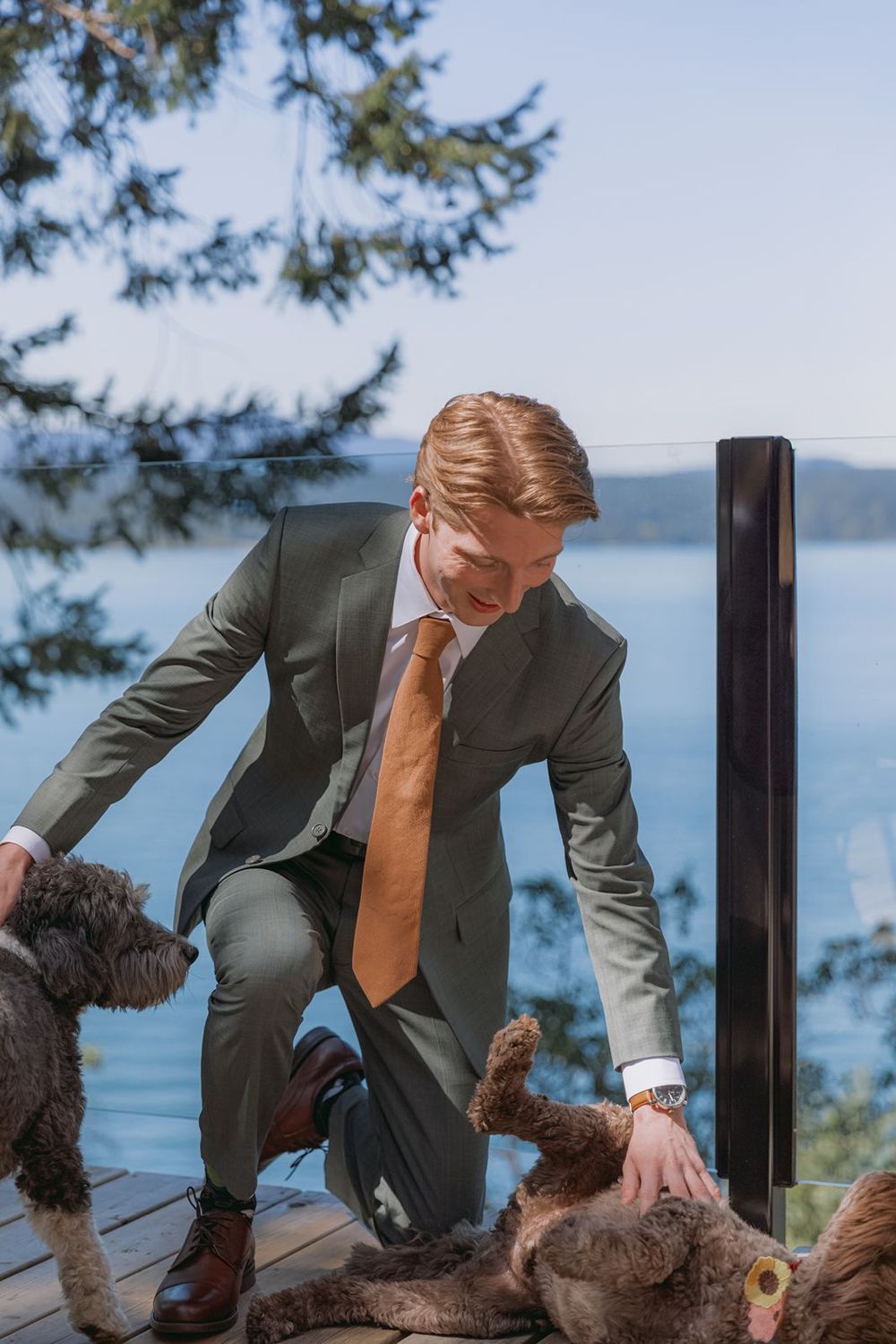 A groom in a gray suit and orange tie kneels on a deck to pet two dogs with a lake view behind him.