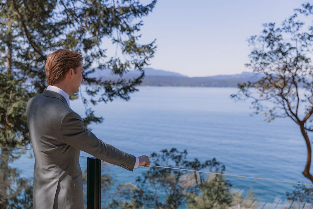 Groom in gray suit gazing at serene blue lake view from balcony railing surrounded by pine trees.
