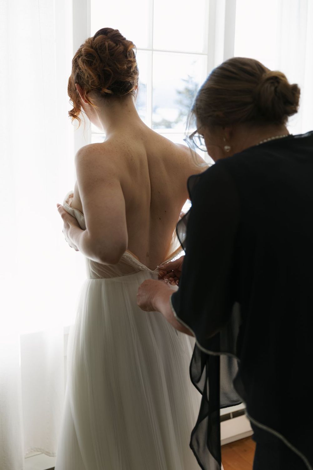 A bride in a backless white wedding gown being helped by a woman near a bright window.