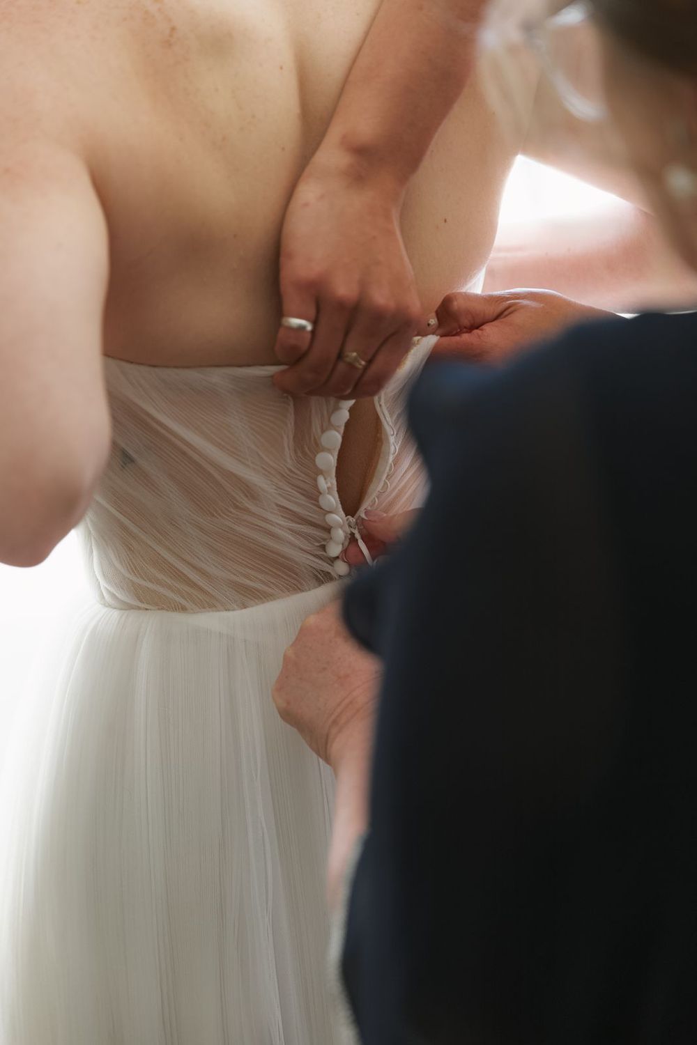Bride getting wedding dress buttoned up the back by a helper during bridal preparations.