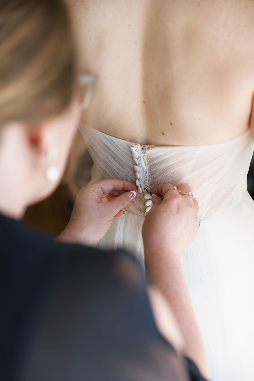 Bridesmaid fastening delicate buttons on the back of a white wedding dress during bridal preparations.