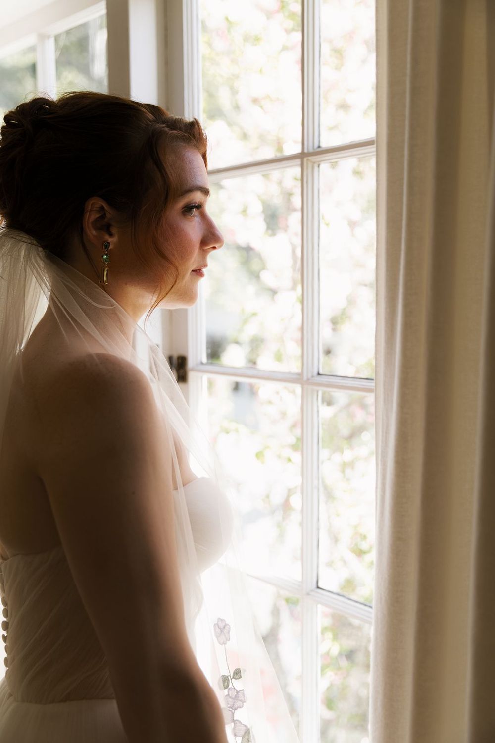 Beautiful bride in white gown and veil with green drop earrings gazing thoughtfully out a sunlit window.