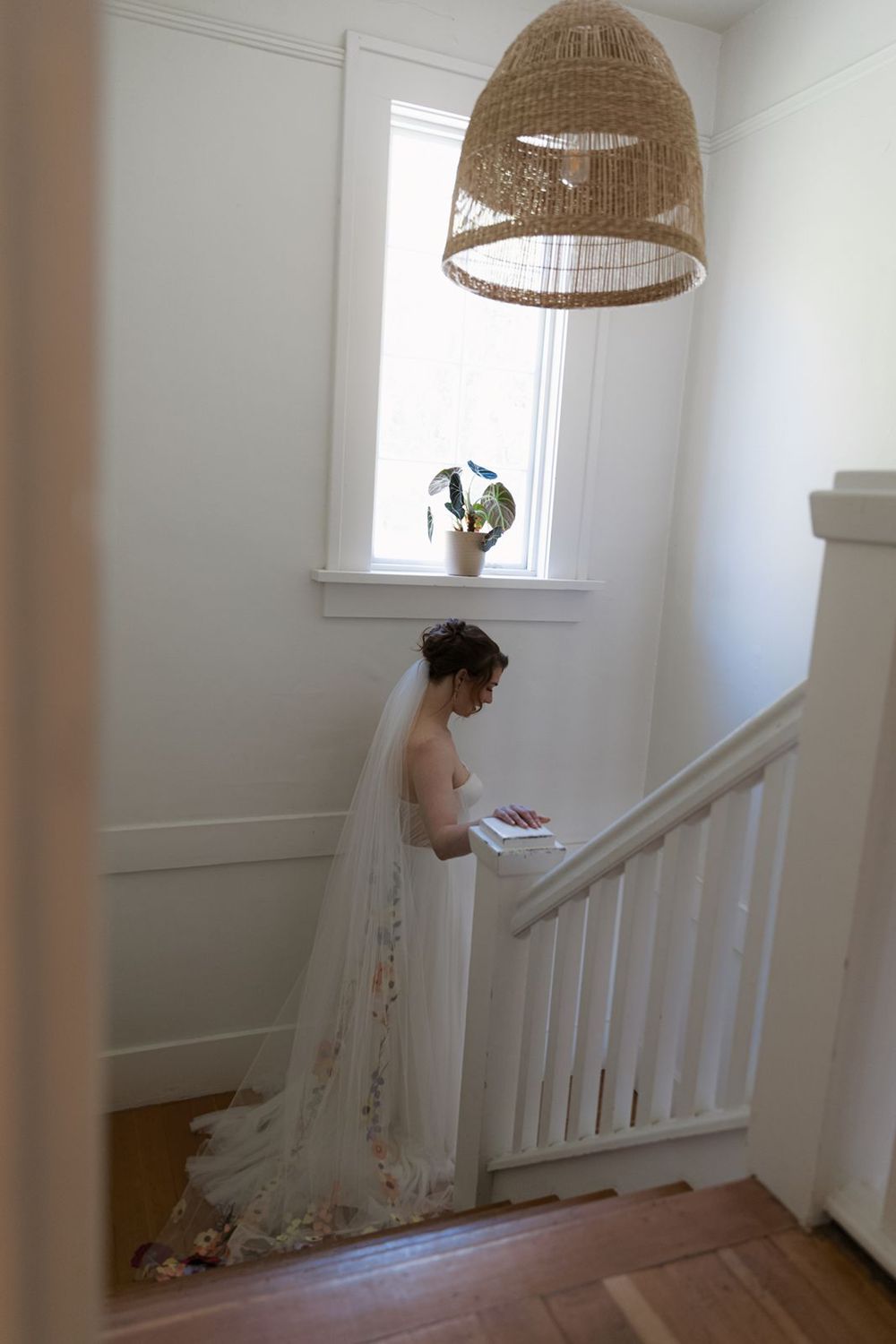 Bride in a floral wedding gown with long veil reading near a staircase window in a bright white home.
