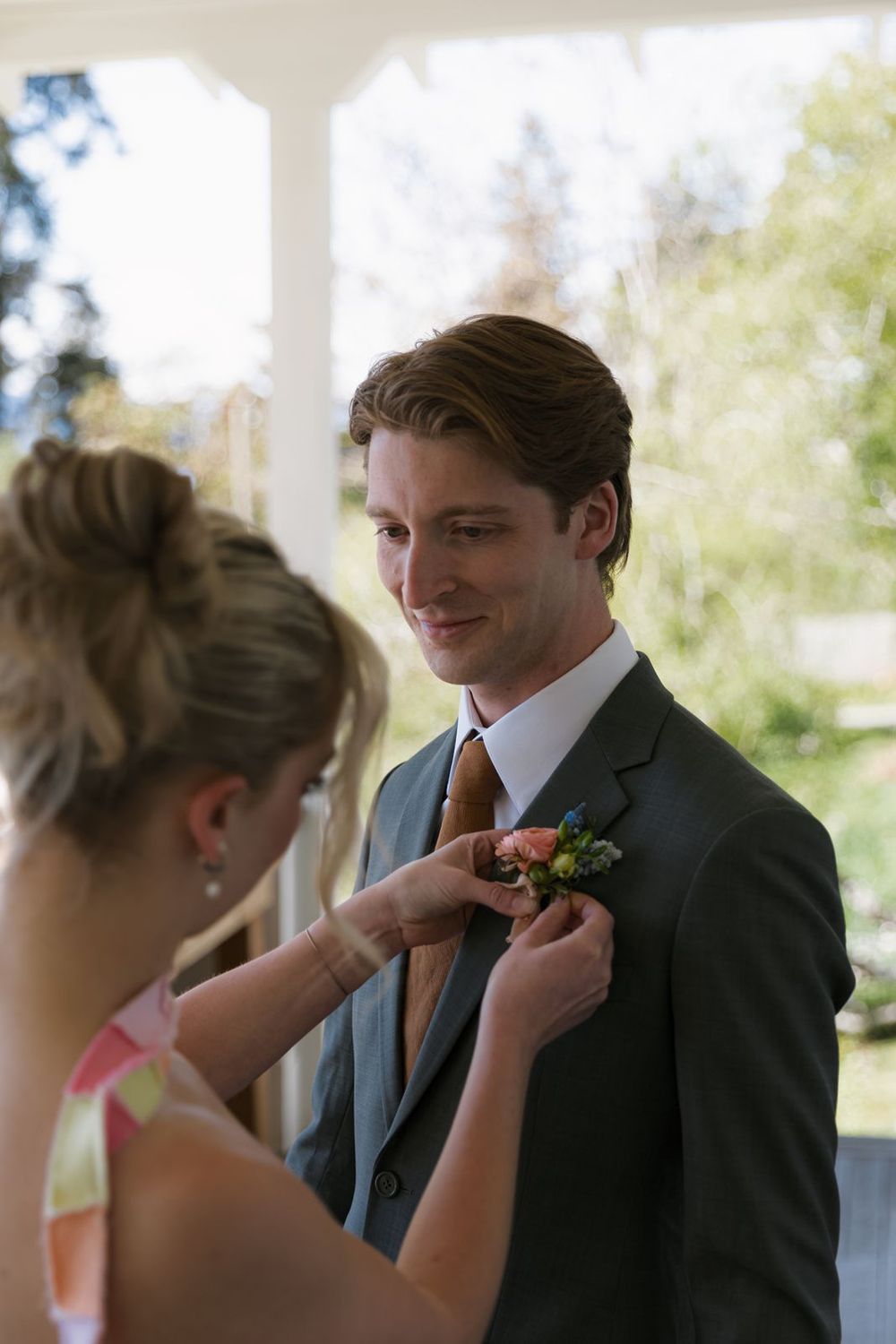 A groomsman smiles as a woman pins a floral boutonniere to his grey suit jacket at an outdoor wedding.