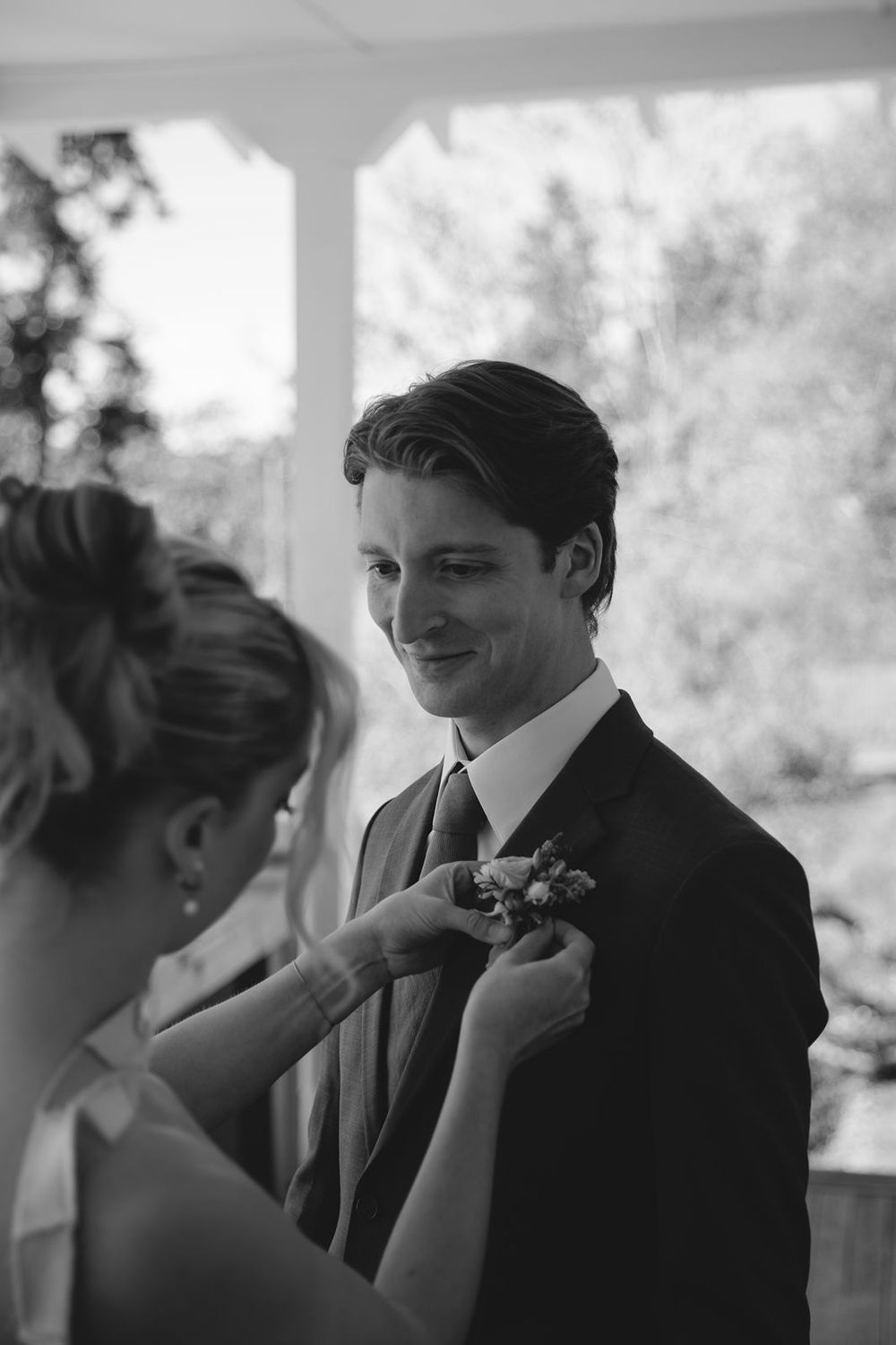 Black and white photo of a groom smiling as a woman pins a boutonniere to his suit lapel on his wedding day.