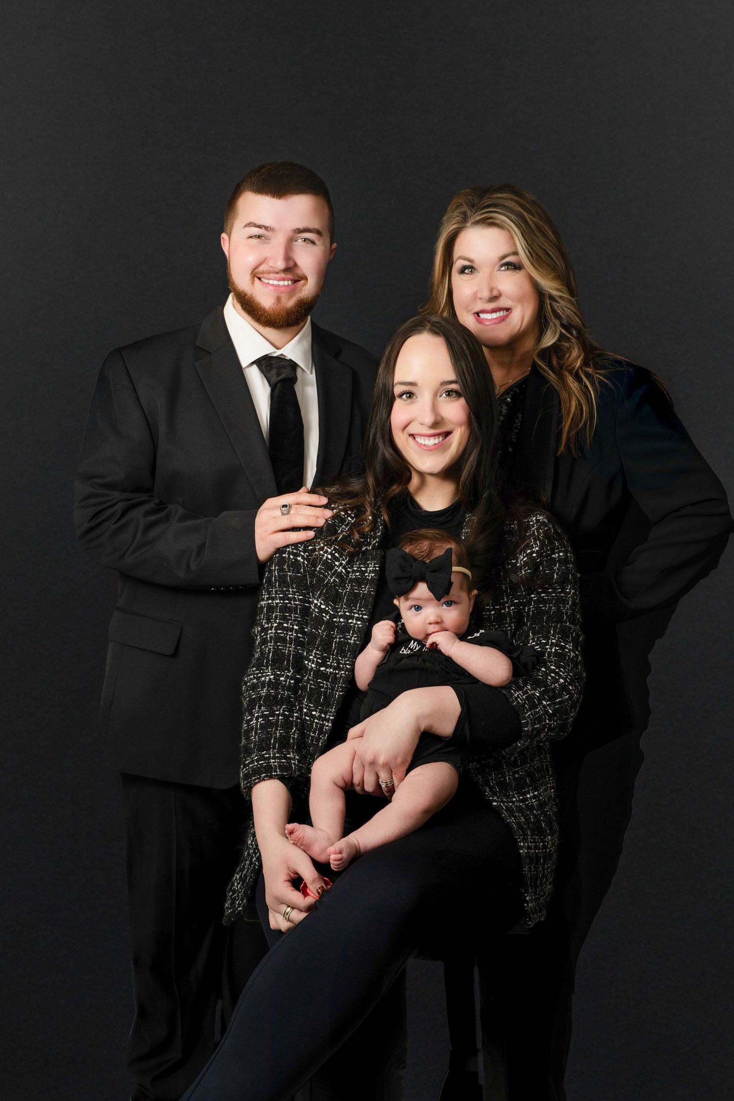 Family portrait of three adults and a baby in black outfits posed against a dark studio background.