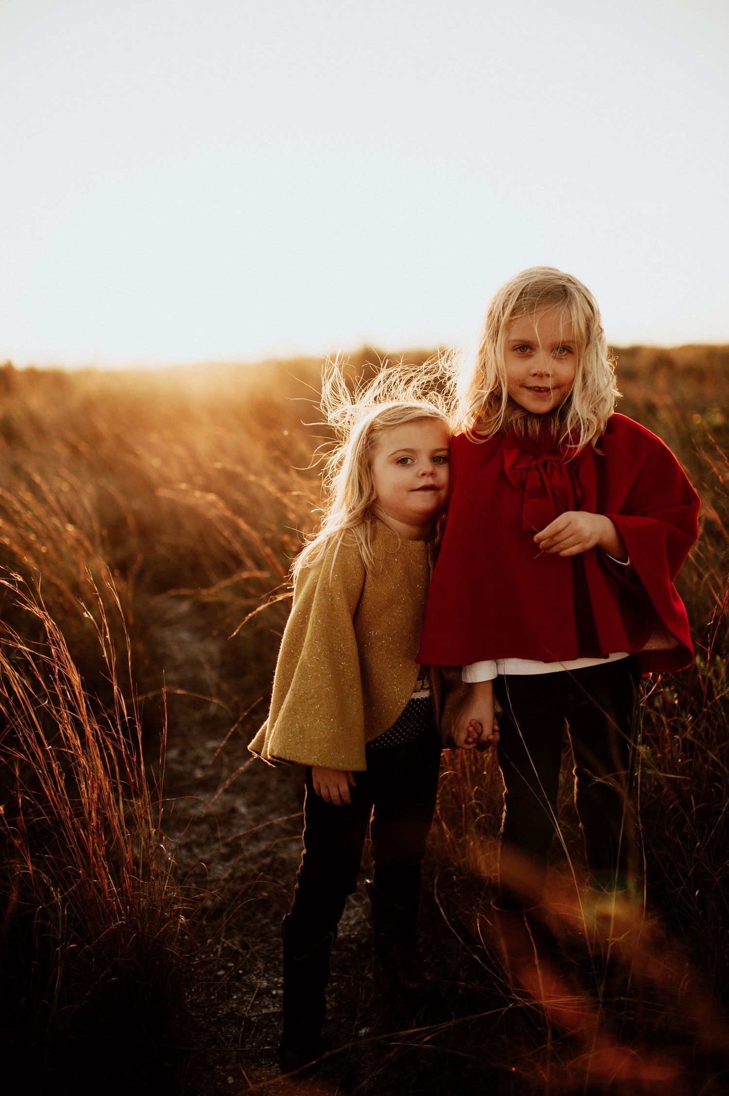 Two young blonde girls in autumn outfits stand in a golden field at sunset, one in red cape, one in mustard sweater.