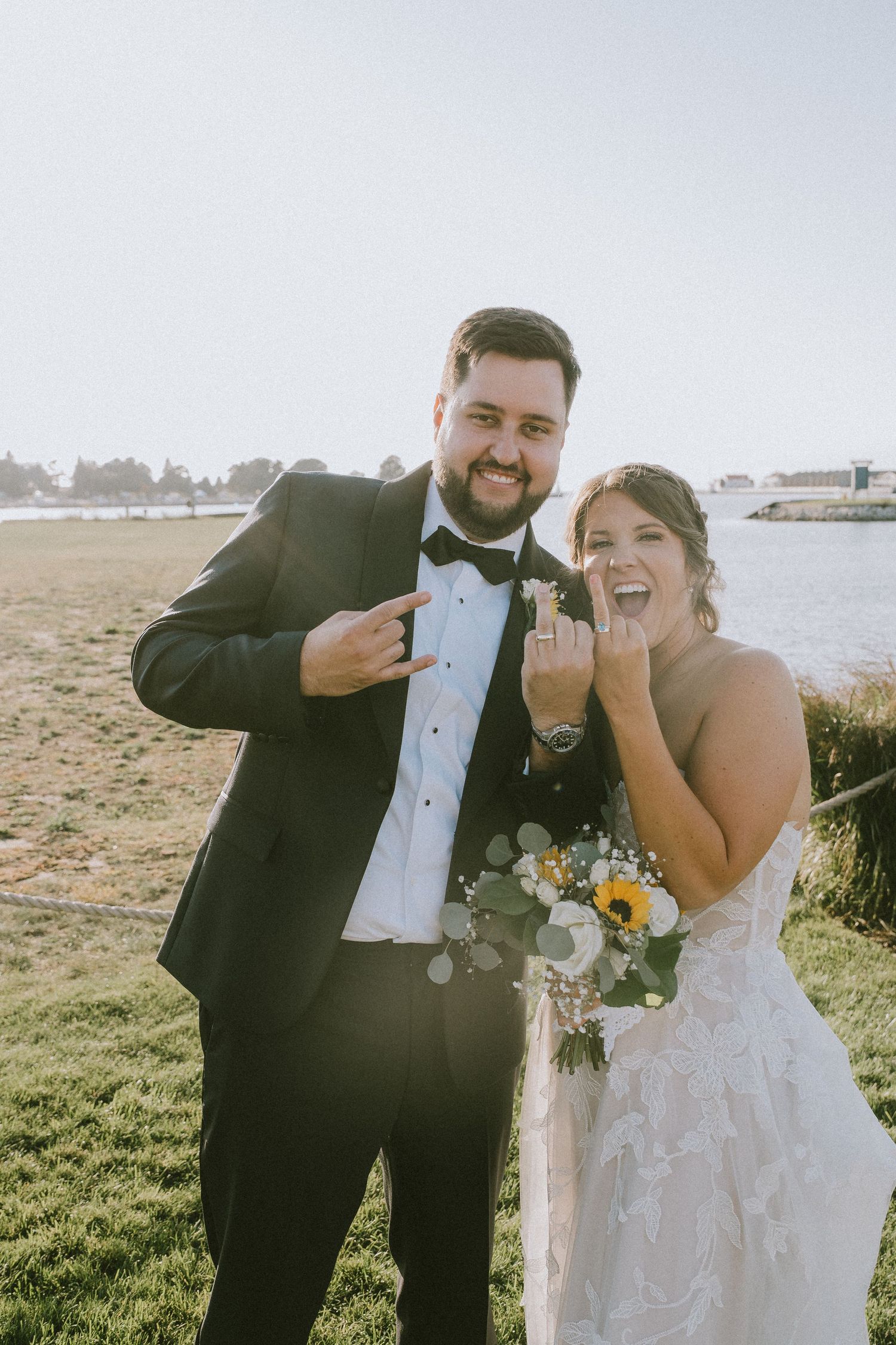 A joyful bride and groom show off their wedding rings outdoors near a waterfront, dressed in a tuxedo and lace gown.