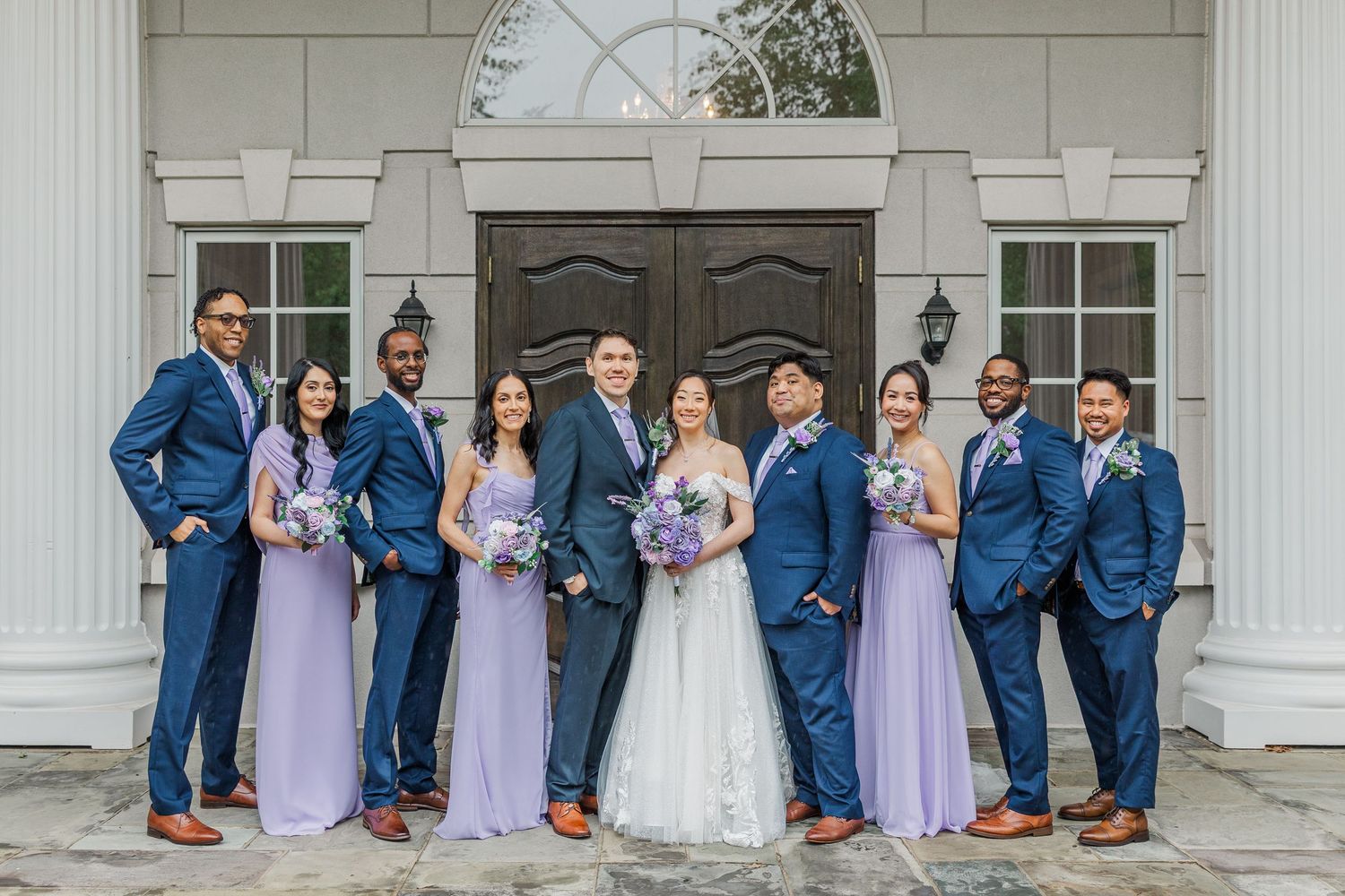 Wedding party in lavender bridesmaid dresses and navy suits posing before elegant white venue doors.