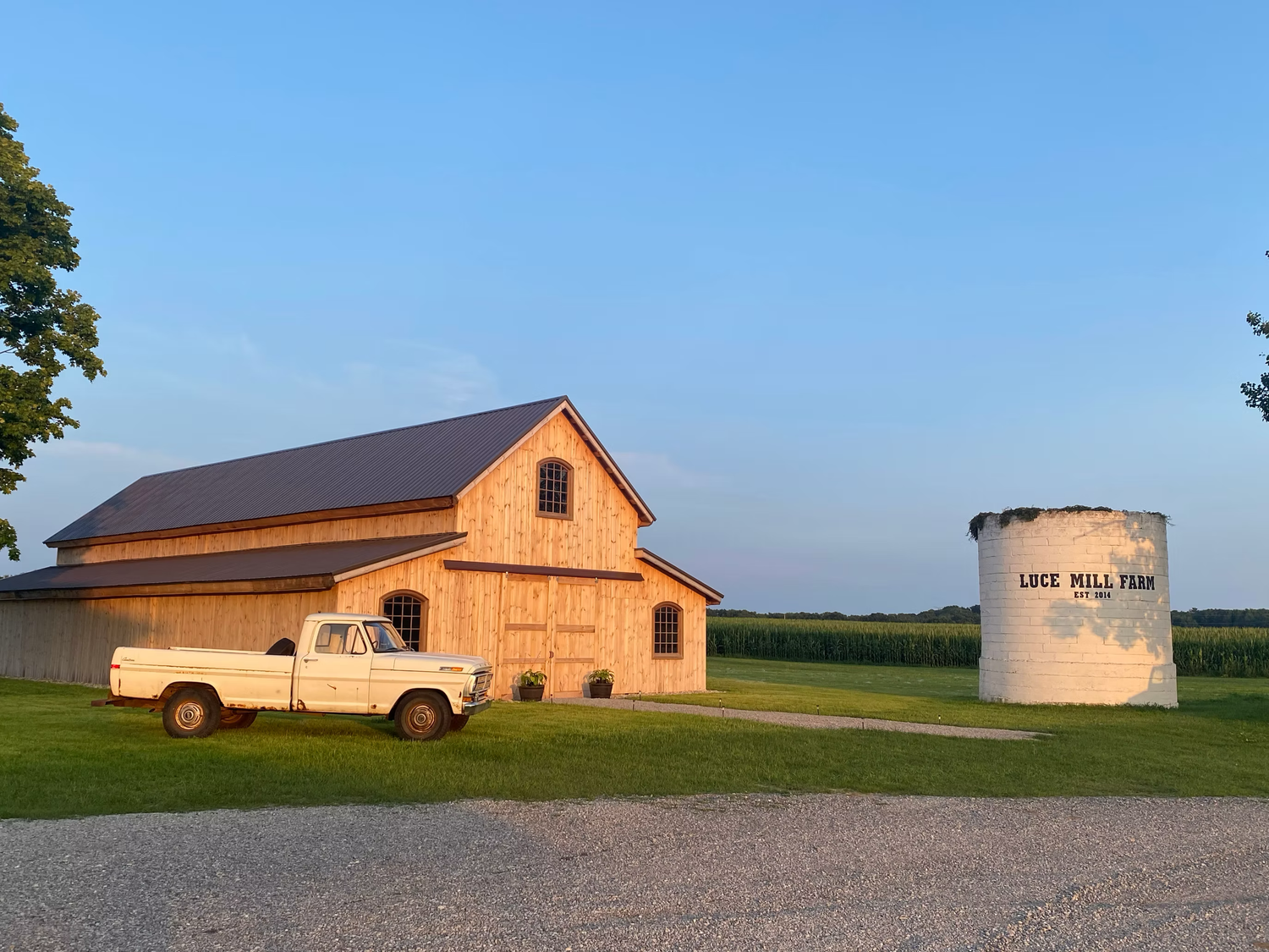 A rustic wooden barn and grain silo at Luce Hill Farm during golden hour, with a white pickup truck parked on a gravel drive.
