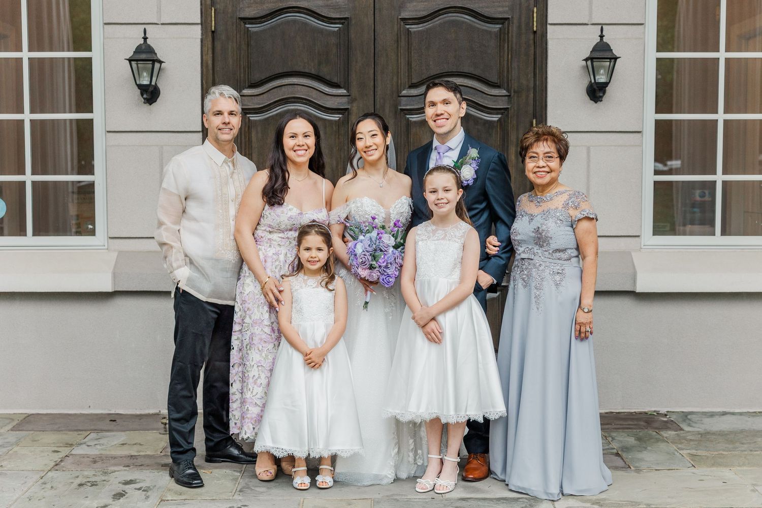 Wedding party poses outdoors, bride and groom flanked by family, two flower girls in white dresses, elegant attire.