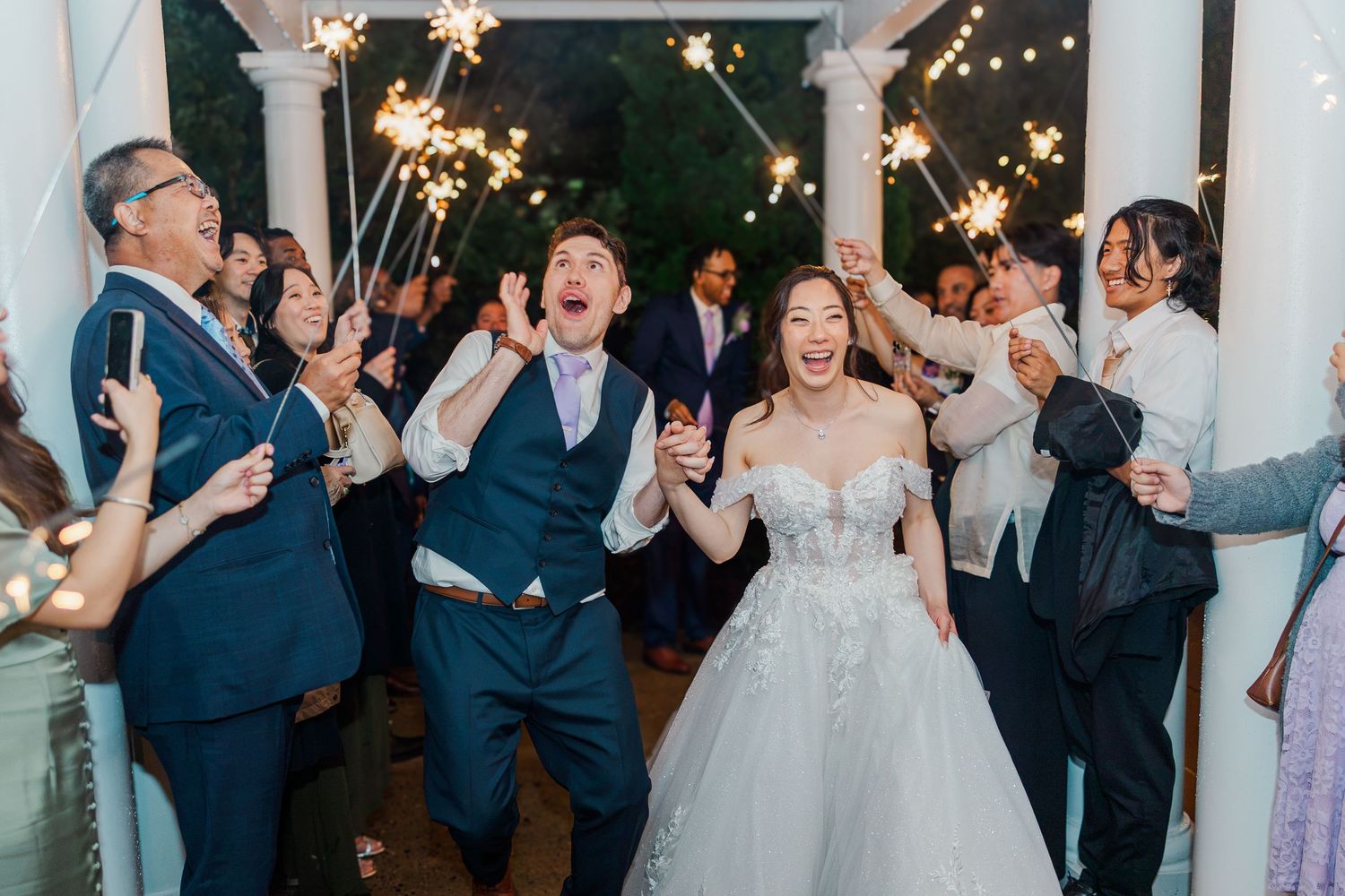 Joyful newlyweds walk through sparkler tunnel surrounded by cheering guests at elegant outdoor wedding reception.