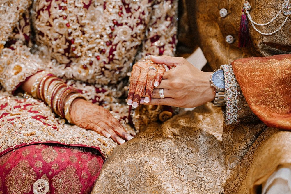Close-up of Indian bride and groom hands adorned with henna, gold jewelry, and embroidered wedding attire.