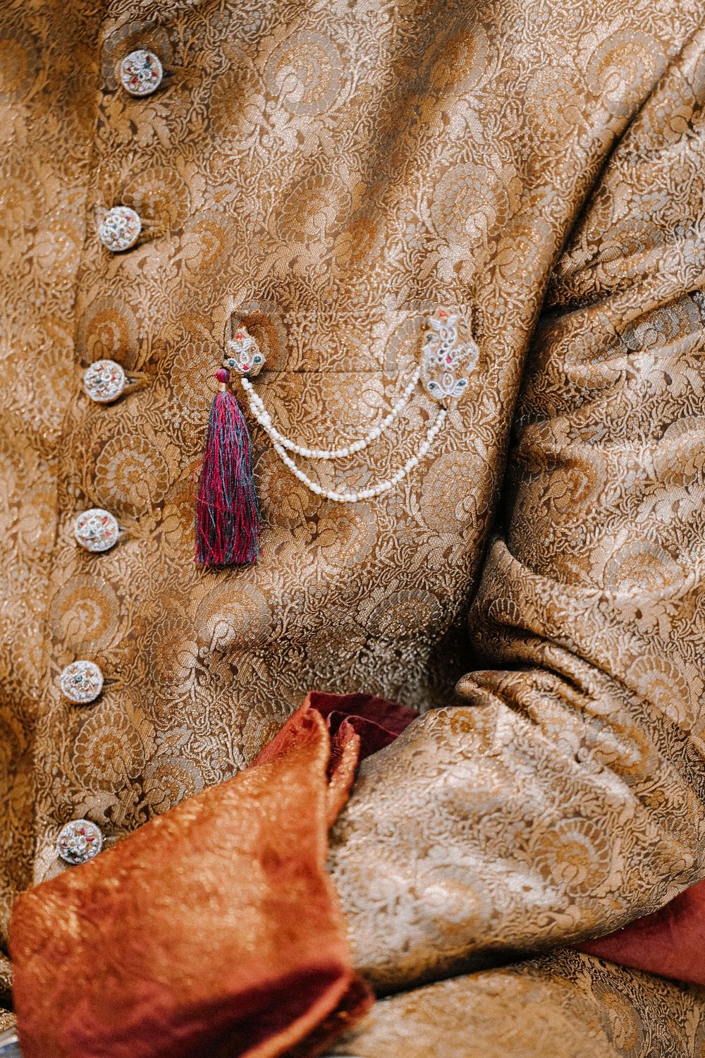Close-up of a golden brocade sherwani with ornate buttons and a decorative tassel brooch pin on the chest pocket.