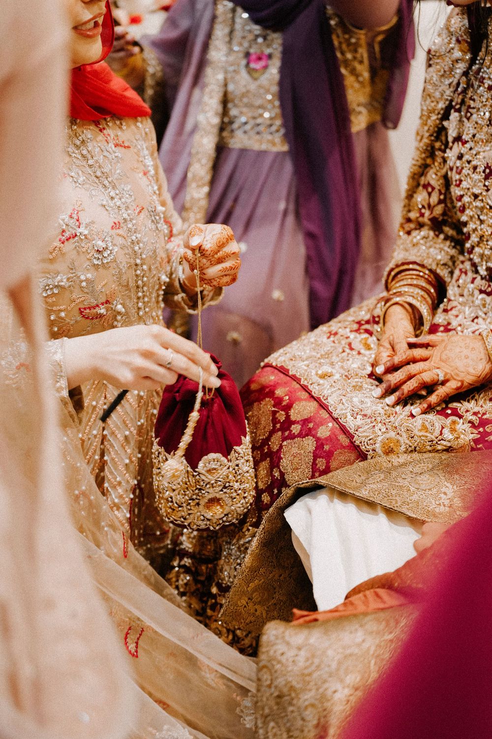 Bride in ornate gold and red lehenga holds decorative golden potli bag at South Asian wedding ceremony.