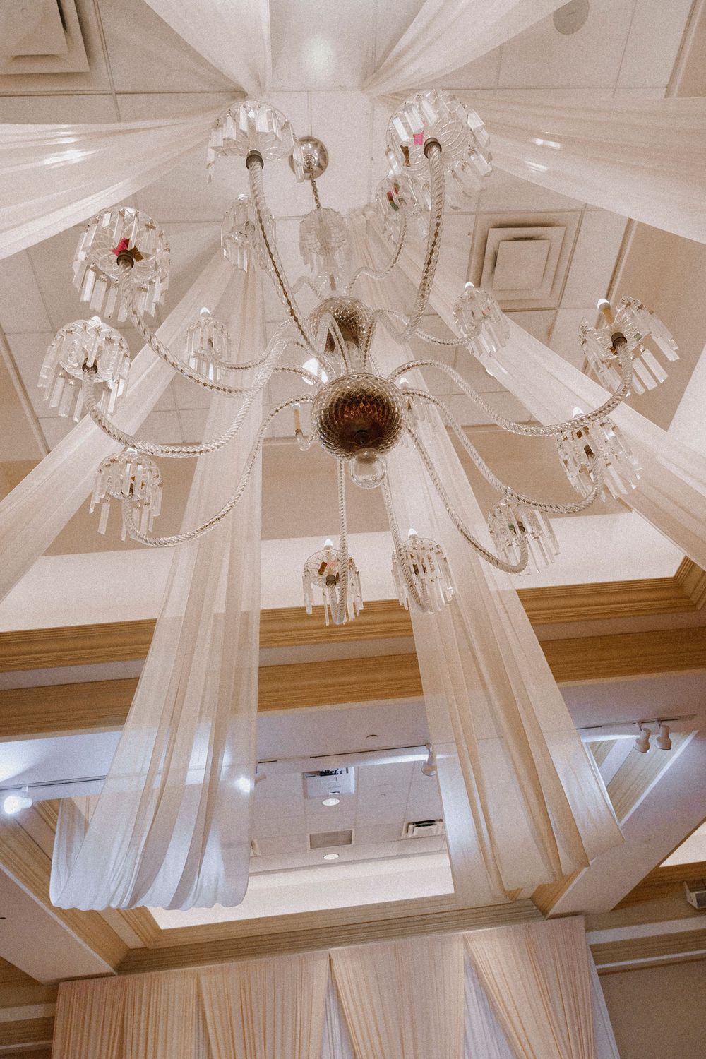Elegant crystal chandelier with white draped fabric hanging from ornate ceiling in luxury ballroom.
