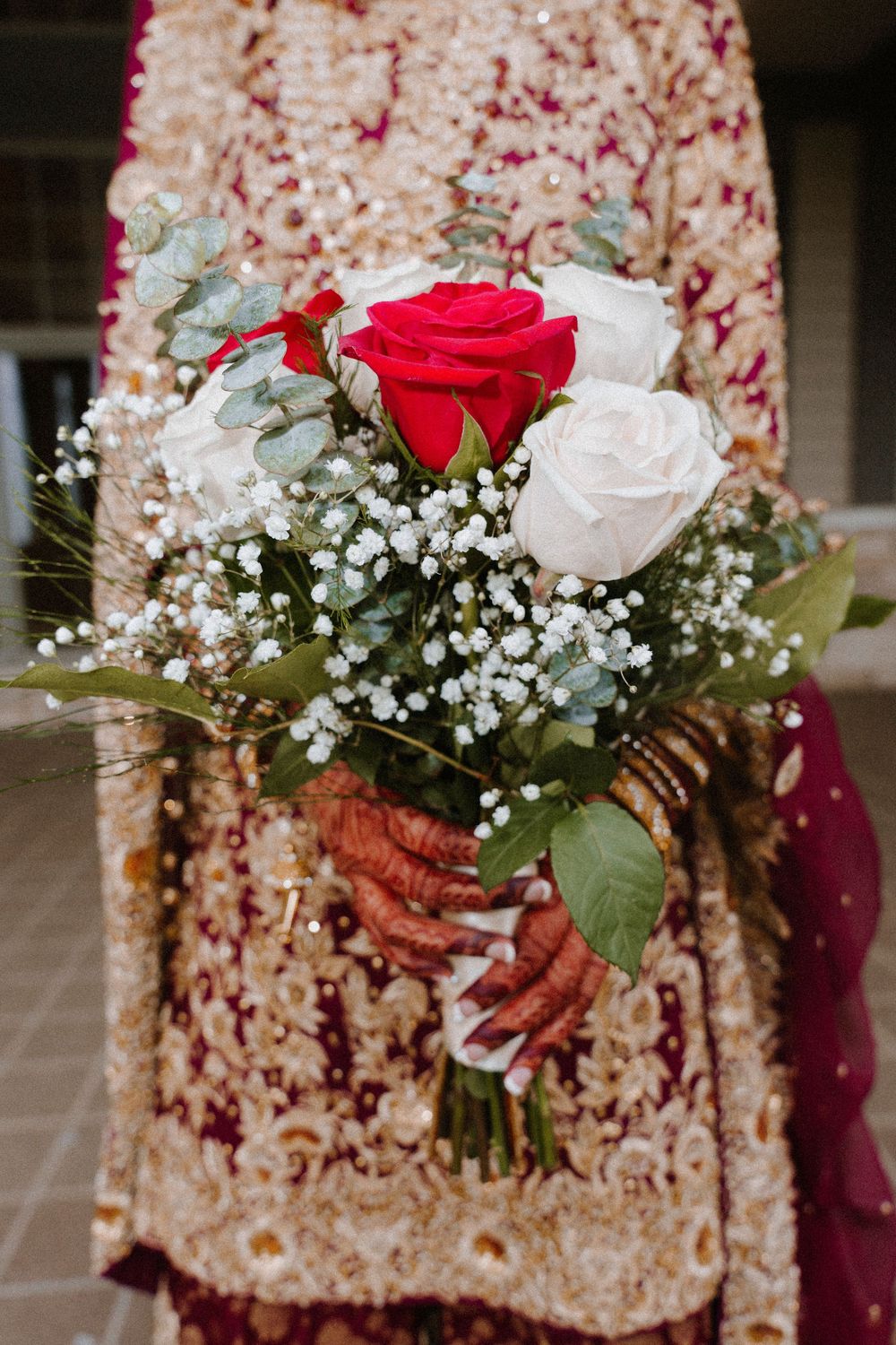 Bride holding a wedding bouquet of red and white roses with baby's breath, wearing ornate embroidered bridal attire.