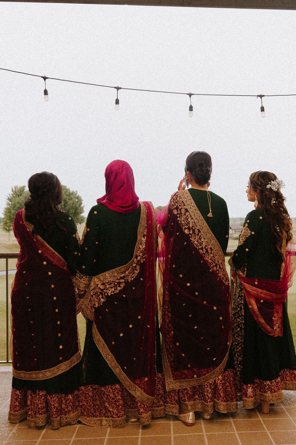 Four women in elegant dark maroon and gold embroidered sarees stand together on a balcony, viewed from behind.