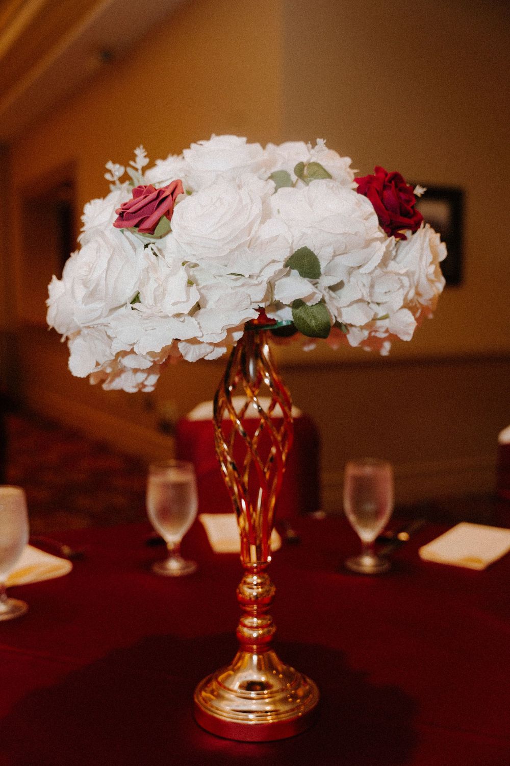 Elegant wedding centerpiece with white and red roses in a gold vase on a red tablecloth with wine glasses.