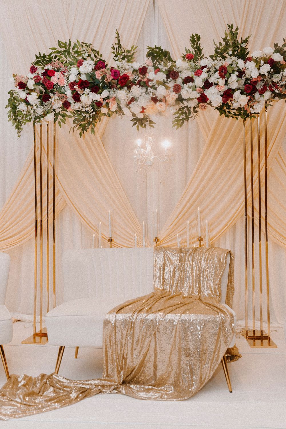 Elegant wedding backdrop with gold stands, floral arch of red and white roses, draped cream fabric, and a gold sequin chair.