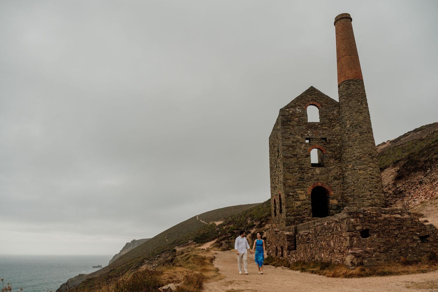 Couple walking near historic Wheal Coates tin mine ruins on Cornwall clifftop with ocean backdrop.