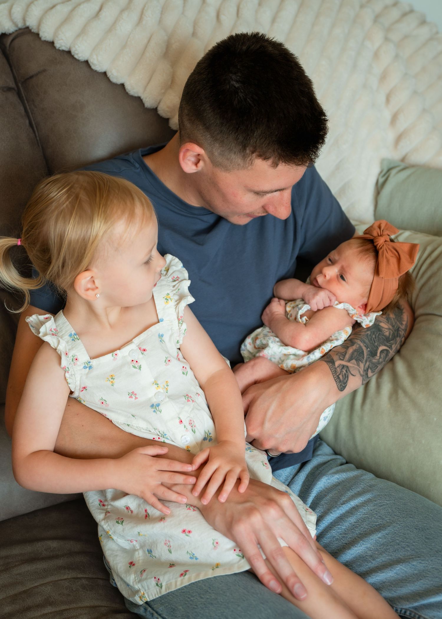 A father sits on a couch cradling a newborn baby while his young blonde toddler daughter leans in closely beside him.
