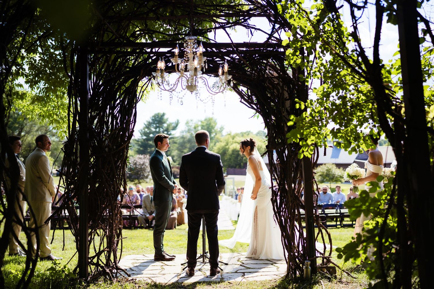 Outdoor wedding ceremony under rustic twig arch with chandelier, bride and groom facing officiant in lush garden setting.