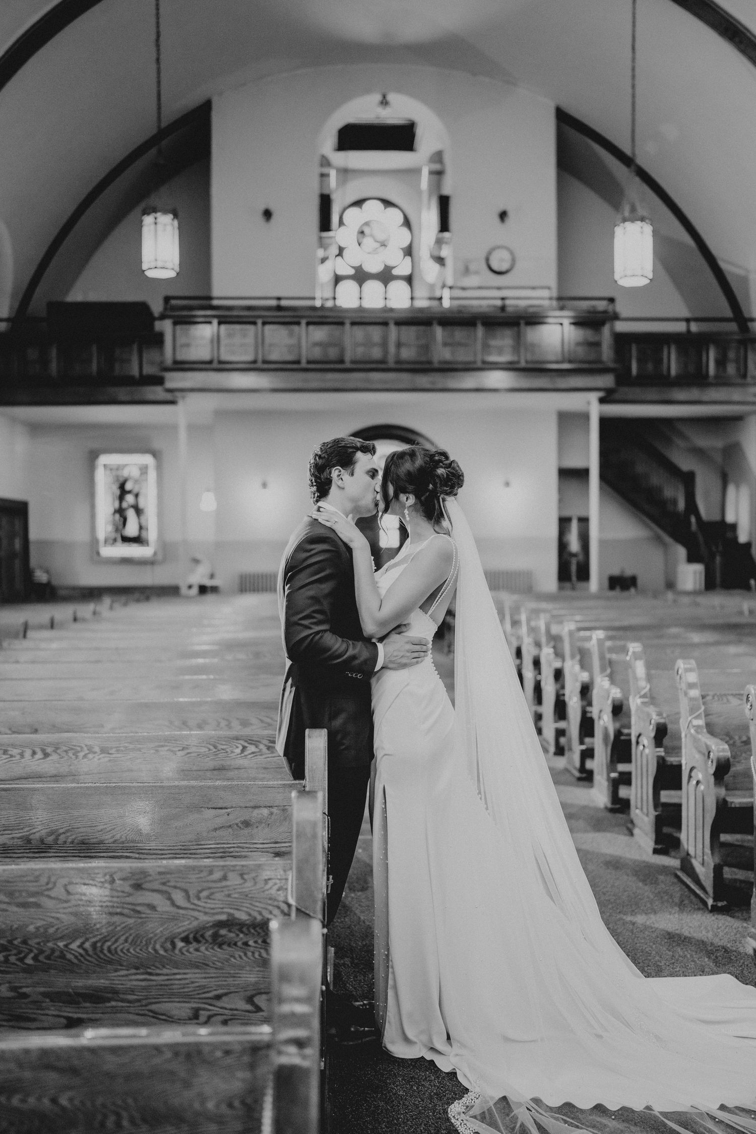 Black and white photo of a bride and groom sharing a kiss in a church aisle, with pews and arched balcony behind them.