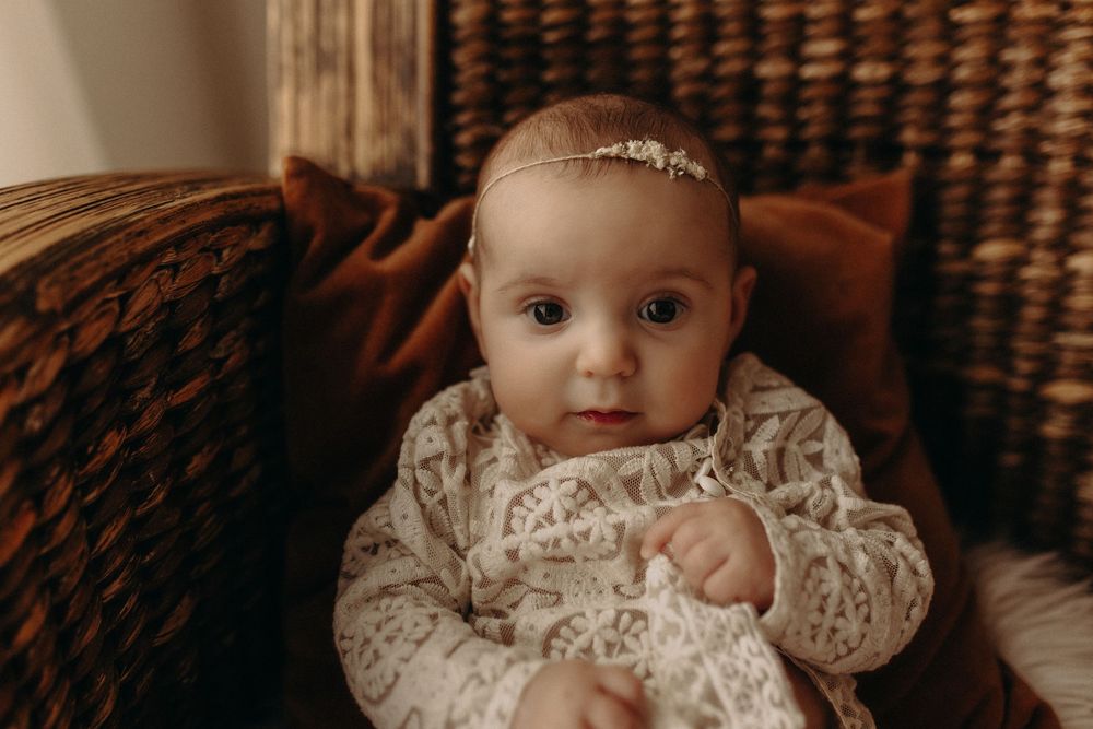 Adorable baby girl in lace dress and floral headband sitting in wicker chair with warm brown tones.
