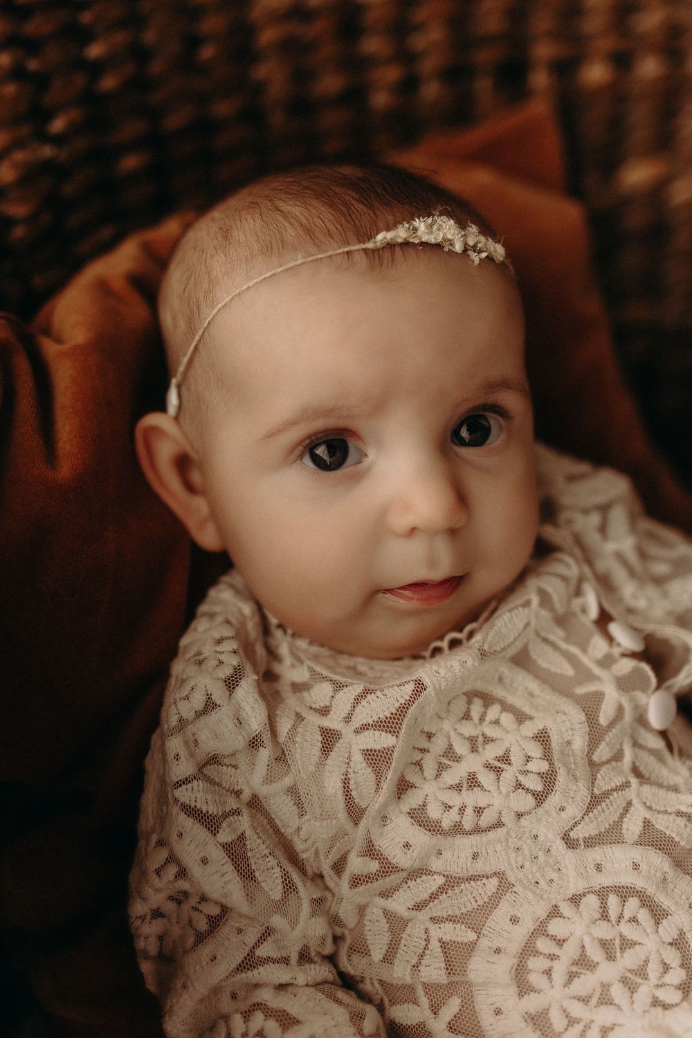 Adorable baby girl in lace dress and delicate headband resting in a wicker basket with warm brown fabric.