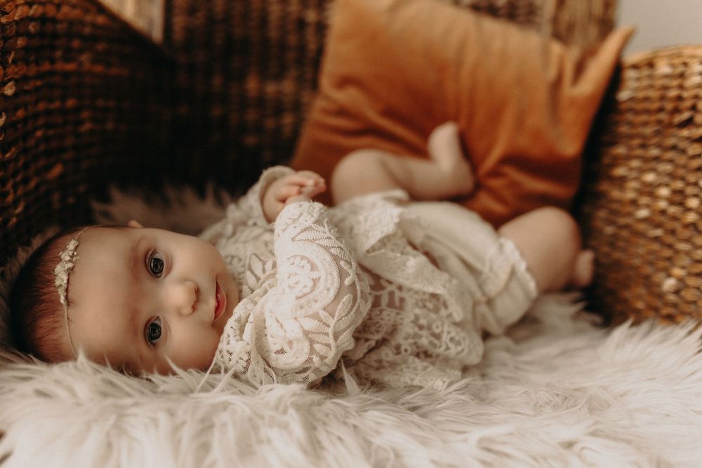 Adorable baby in lace outfit lying on fluffy white rug surrounded by warm-toned pillows and wicker baskets.
