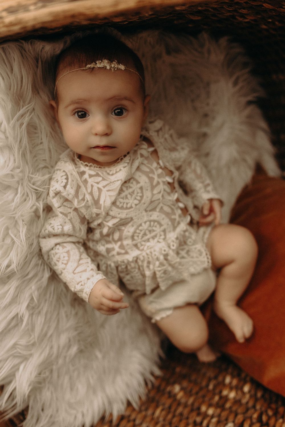 Adorable baby girl in lace romper and floral headband lying on fluffy white fur rug in wicker basket.