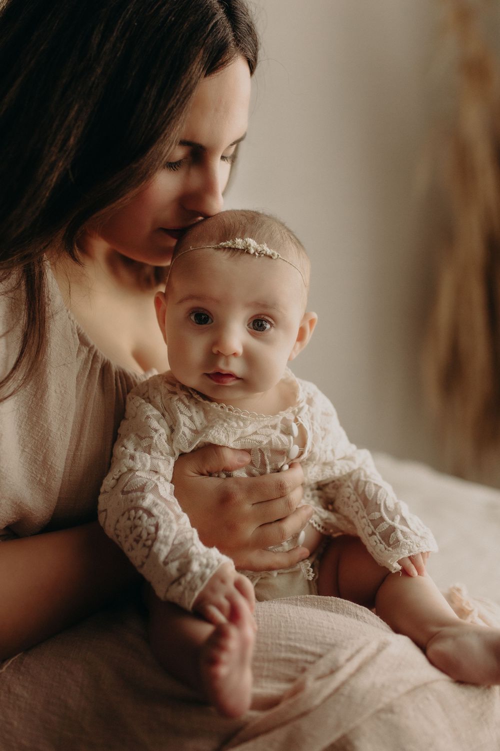 Loving mother kissing baby girl in lace outfit with floral headband during newborn photo session.