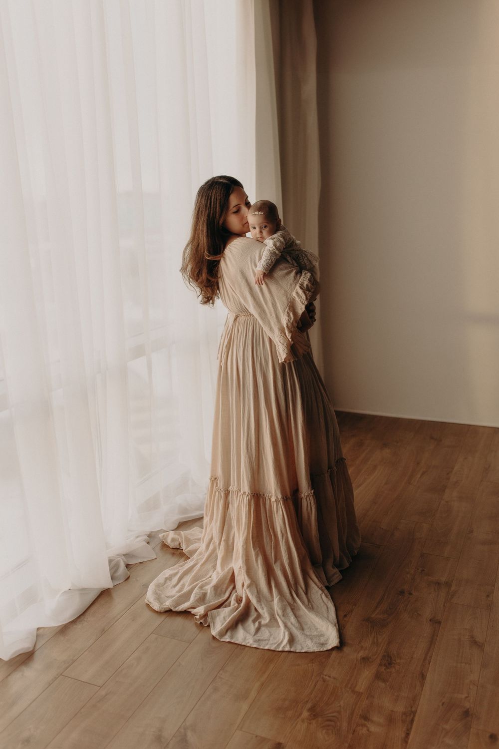 Elegant mother in flowing beige dress holding newborn baby near sheer curtained window in natural light.