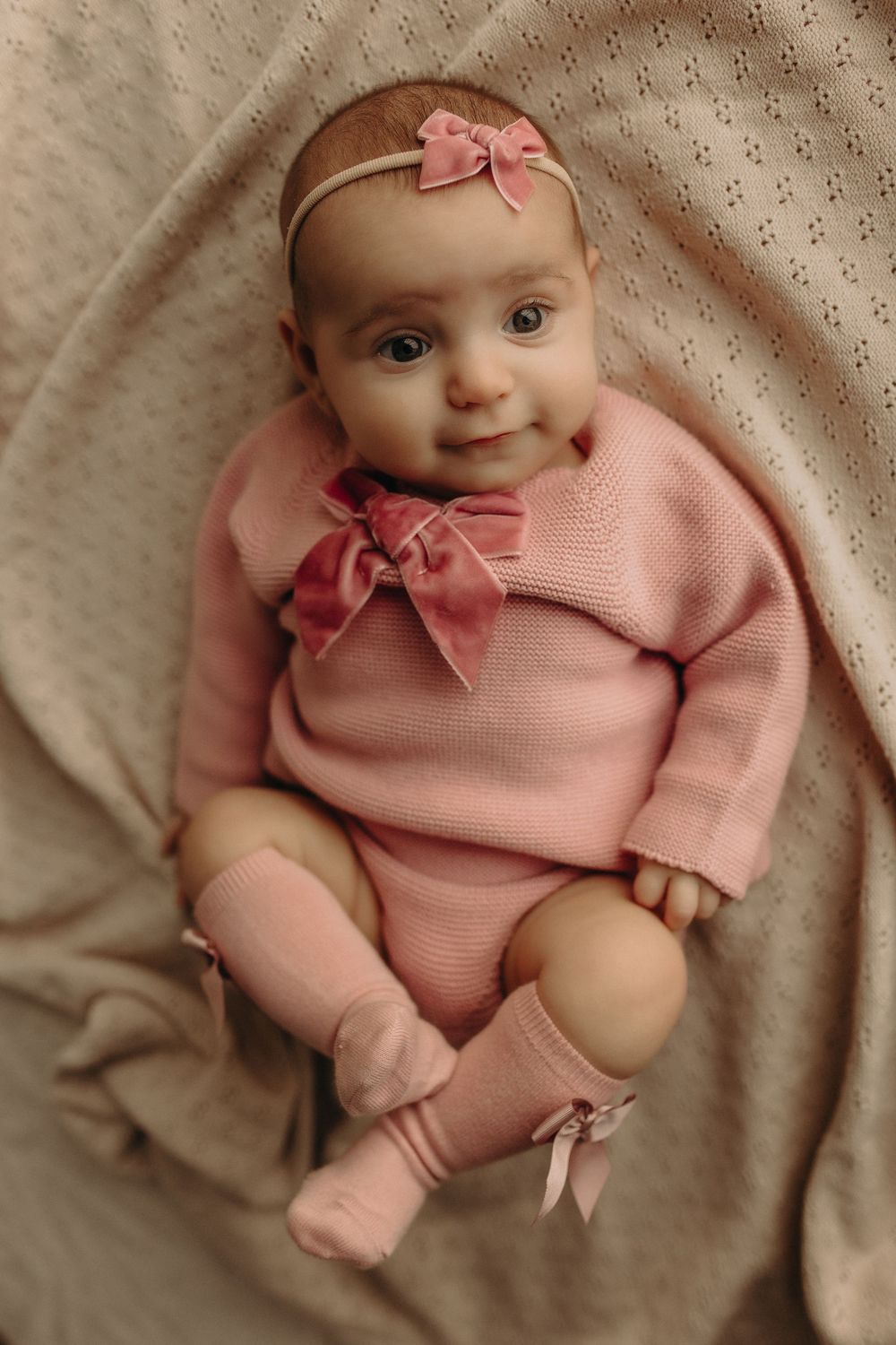 Adorable baby girl in pink knit outfit with bow headband and socks, lying on a beige blanket, smiling sweetly.