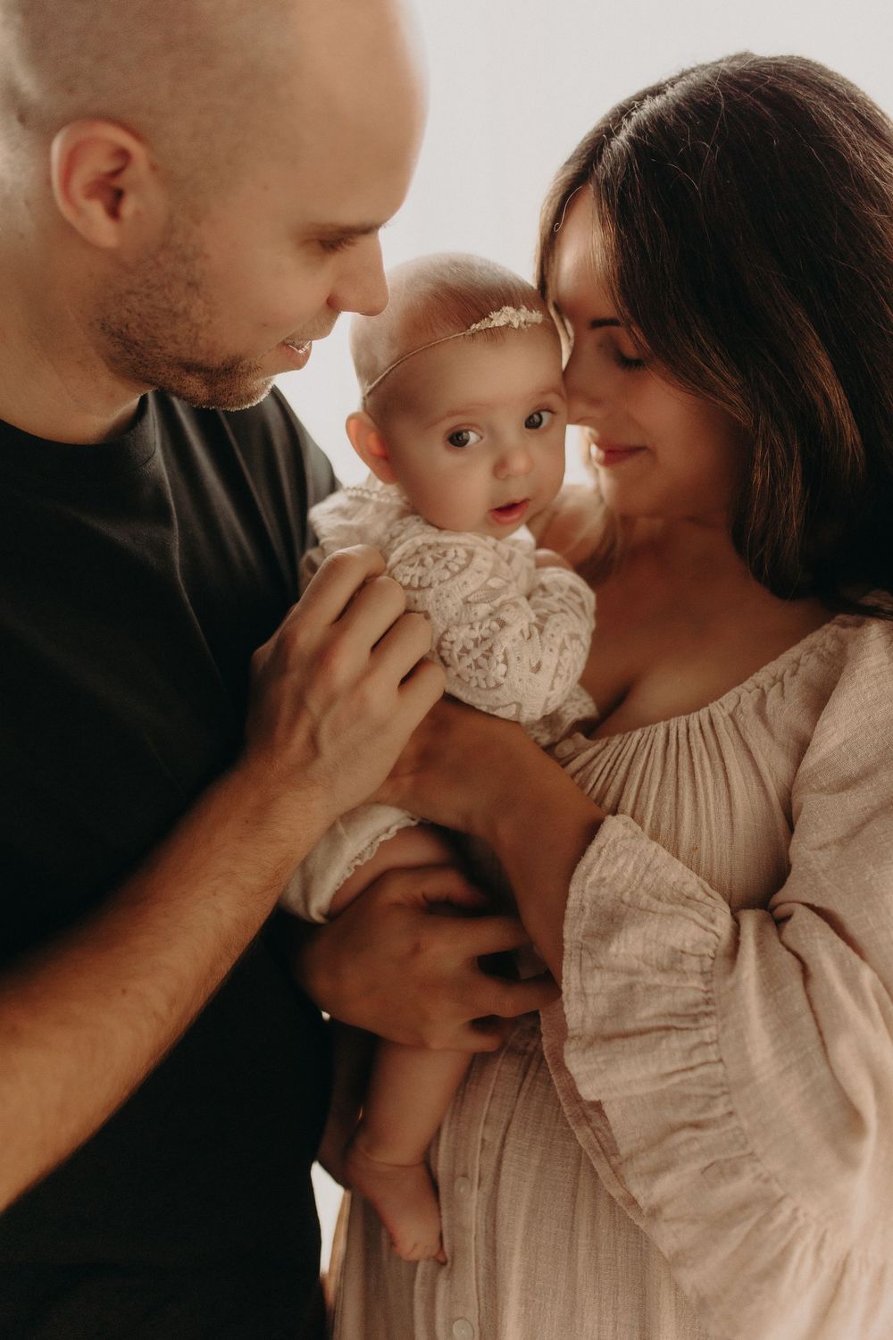 A bald father and brunette mother lovingly hold their baby girl in a white lace headband for a family portrait.