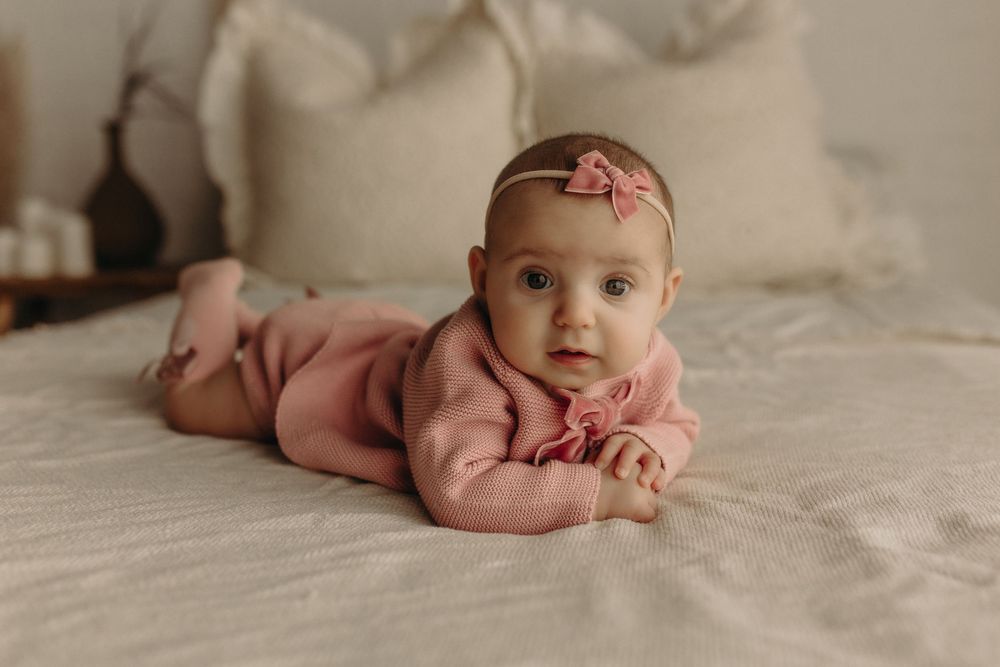 A baby girl in a pink romper and floral headband lies on her tummy on a white bed, looking at the camera.