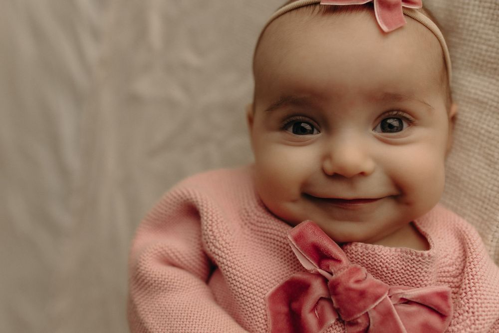 Smiling baby girl in pink knit sweater and bow headband, looking up with bright eyes and a joyful expression.