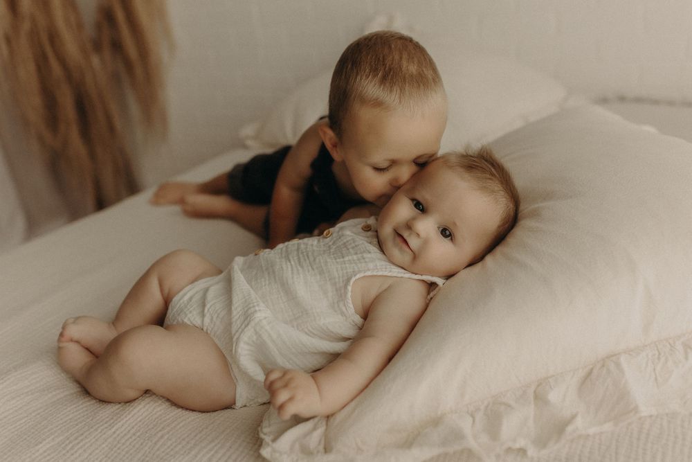 A toddler gently kisses a smiling baby sibling lying on a white bed, capturing a sweet tender moment.