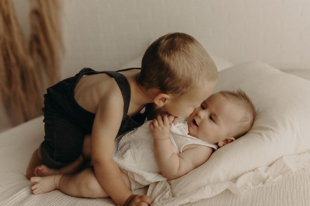 Toddler in black romper gently kissing a smiling baby lying on a white bed.