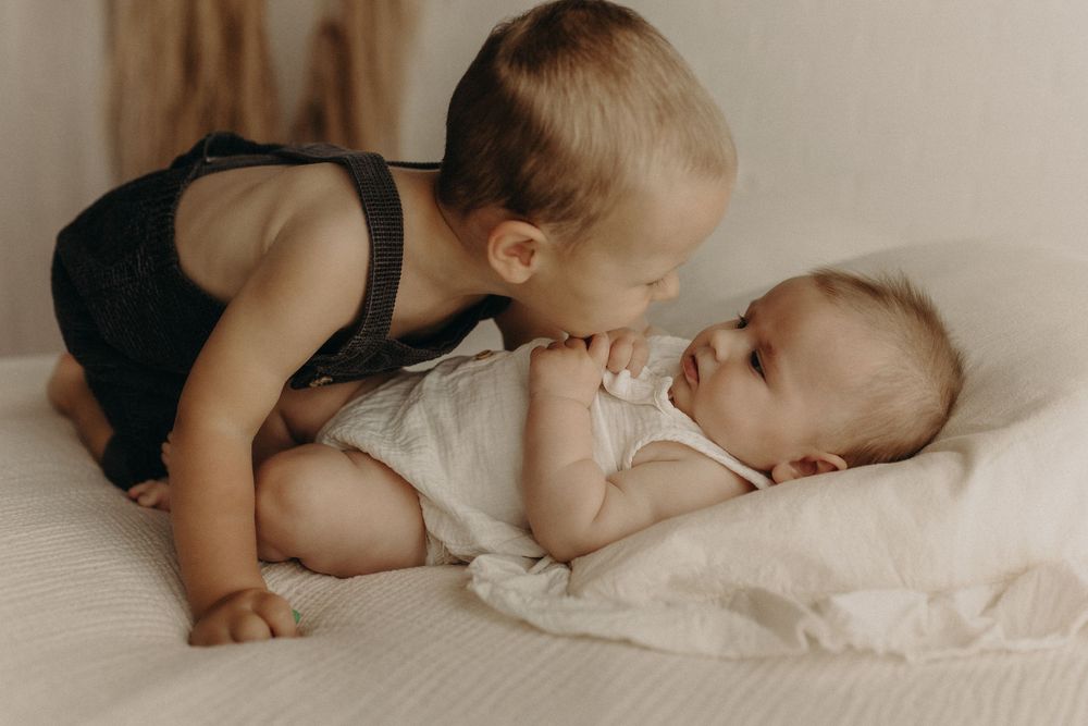 Toddler boy in black romper gently kissing newborn baby sibling lying on white bedding indoors.