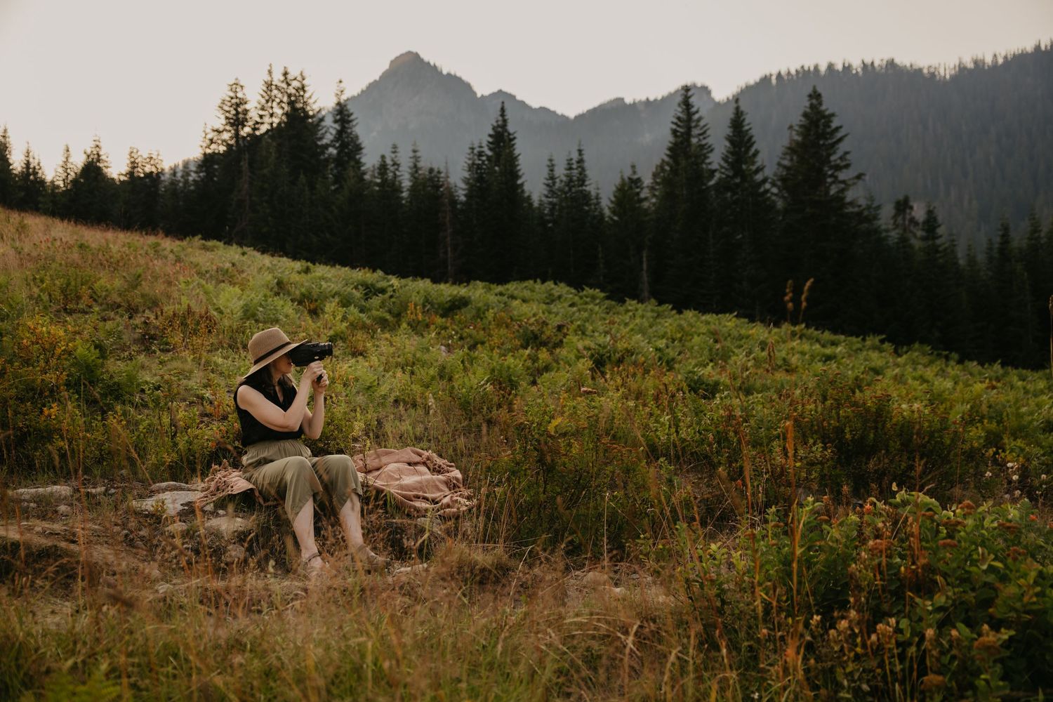 A photographer sits on rocks in a mountain meadow surrounded by evergreen trees, capturing nature with a camera.