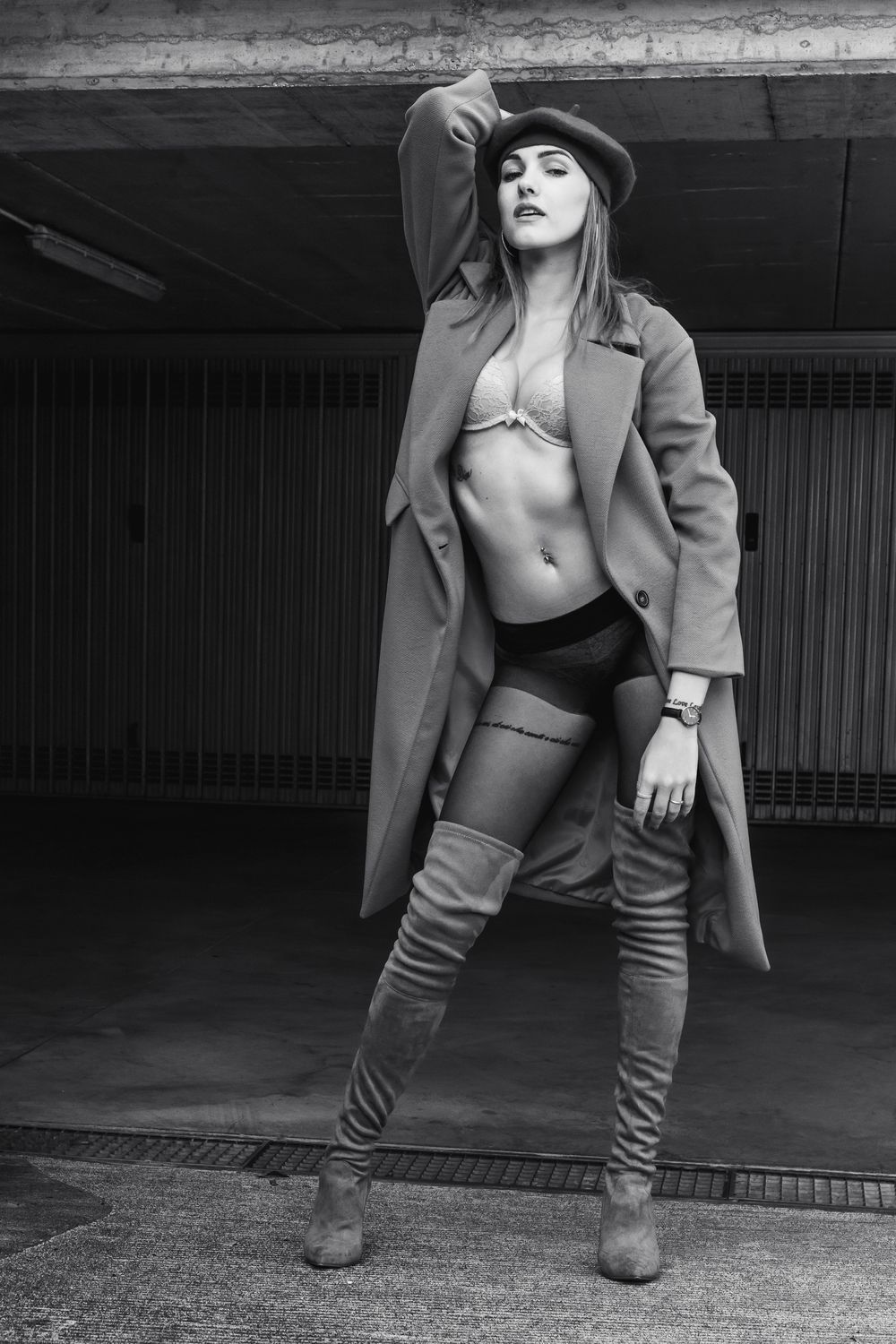 Ritratto glamour in bianco e nero di una modella con cappotto aperto, intimo, basco e stivali alti al ginocchio in una location urbana.