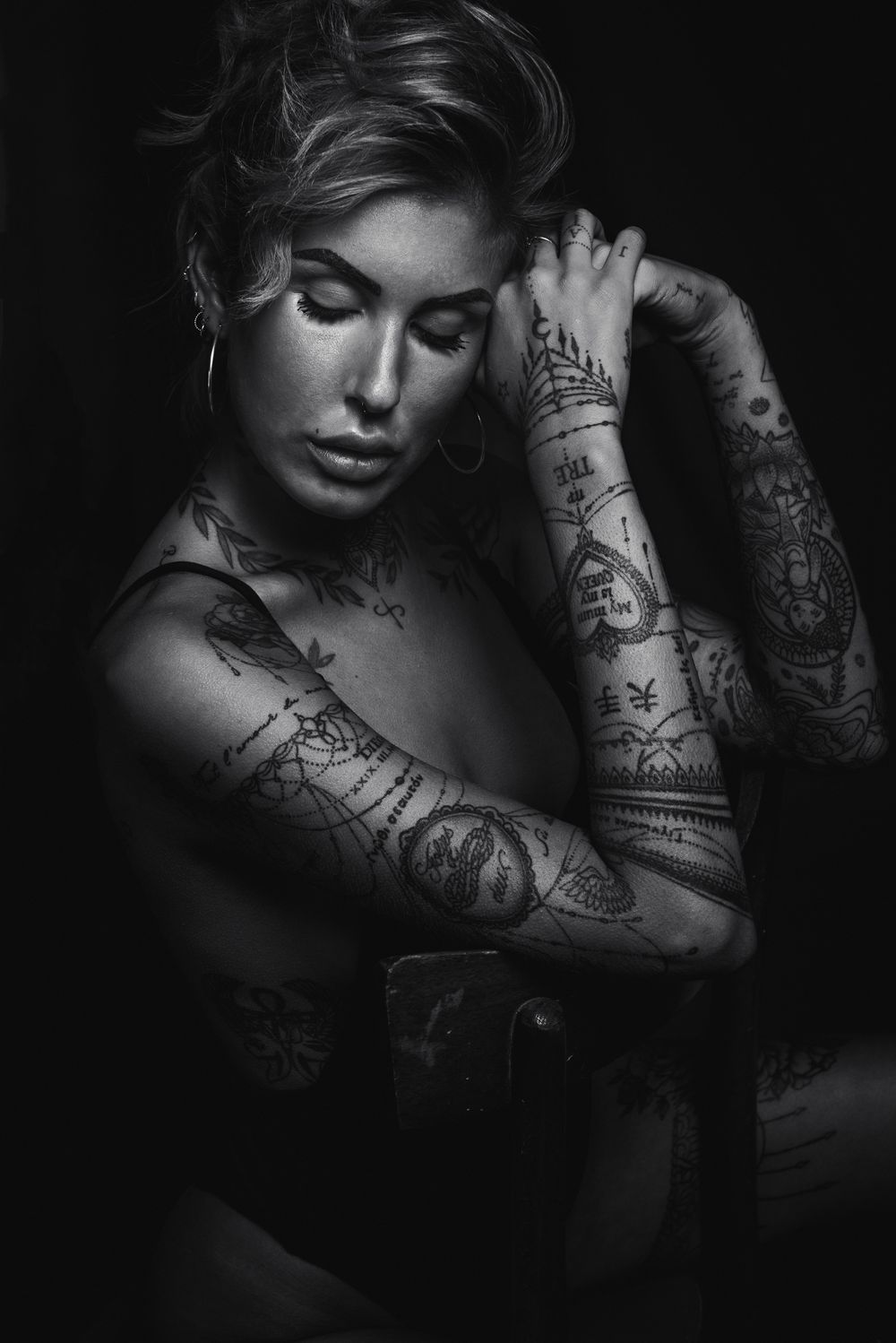 Ritratto glamour intimo e low-key in bianco e nero di una modella tatuata seduta su una sedia, con braccia intrecciate, occhi chiusi e luce