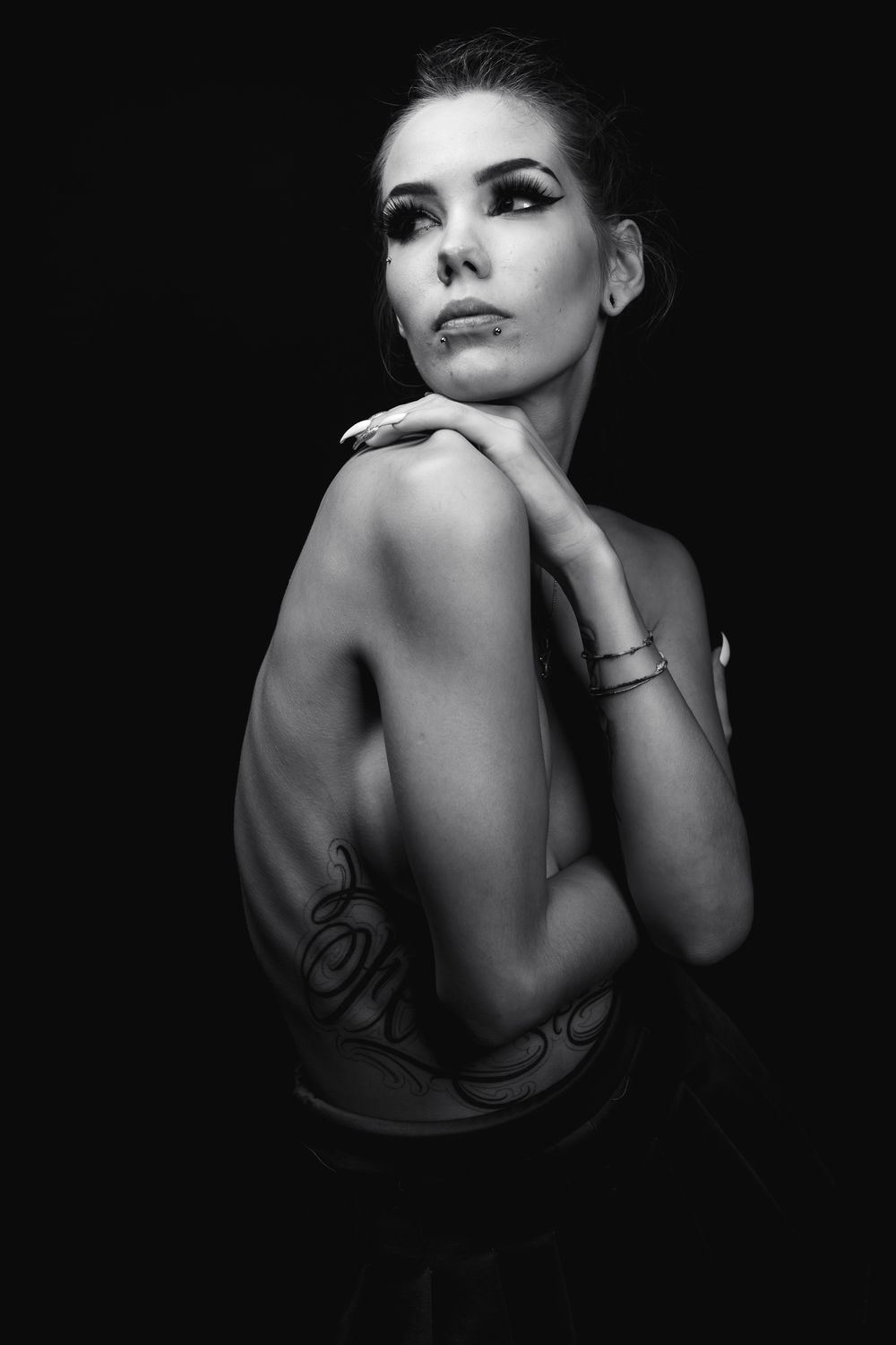 Ritratto glamour intimo e low-key in bianco e nero di una modella tatuata seduta su una sedia, con braccia intrecciate, occhi chiusi e luce