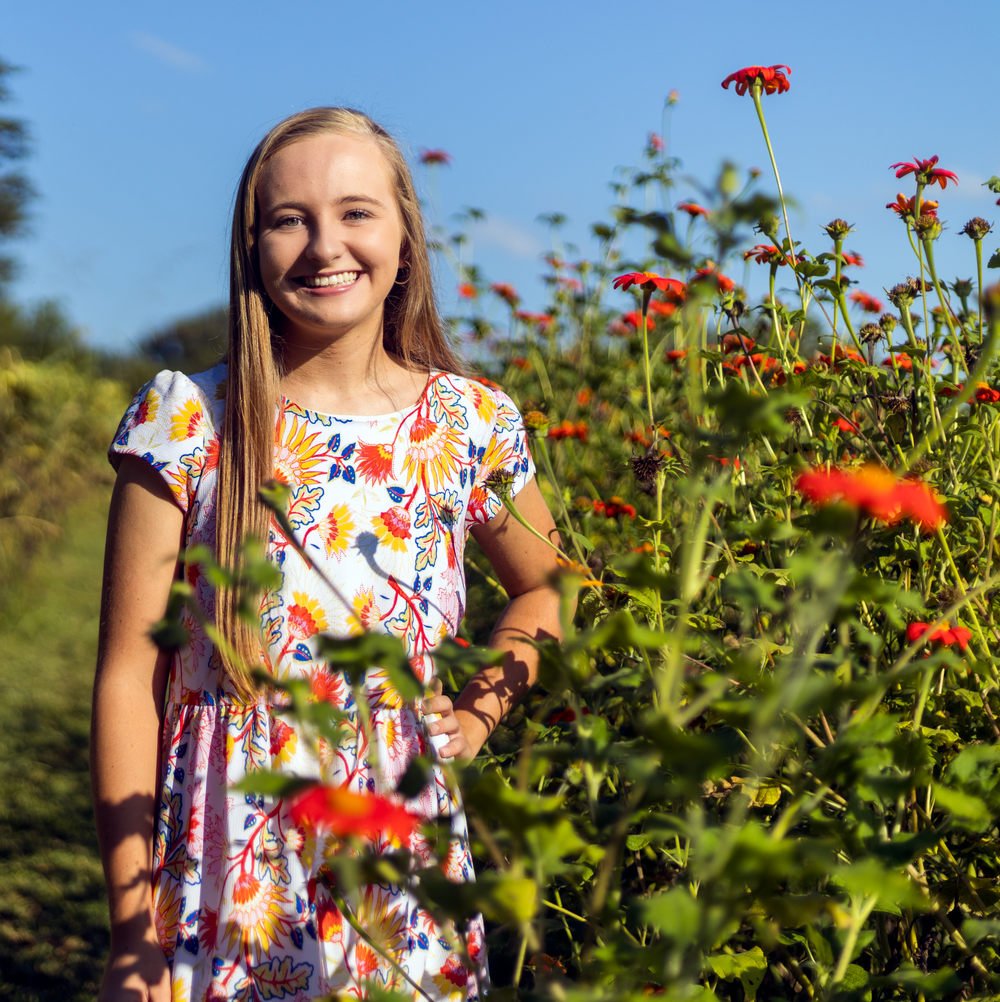 Smiling teenage girl in floral dress standing among vibrant red flowers in a sunny garden.