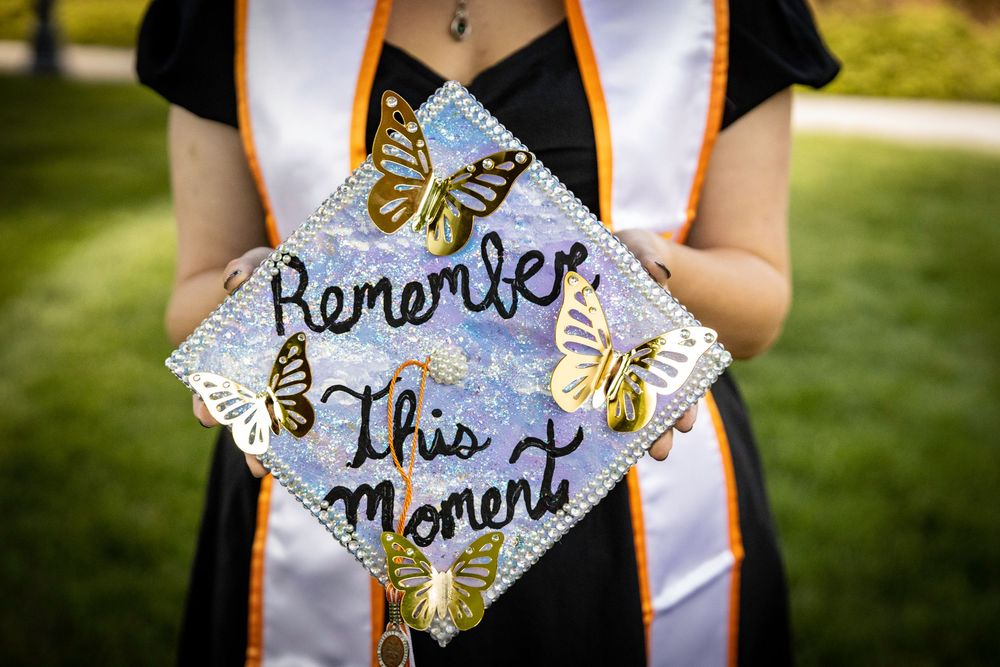 Decorated graduation cap with butterfly designs and cursive text reading Remember This Moment held by graduate in cap and gown.